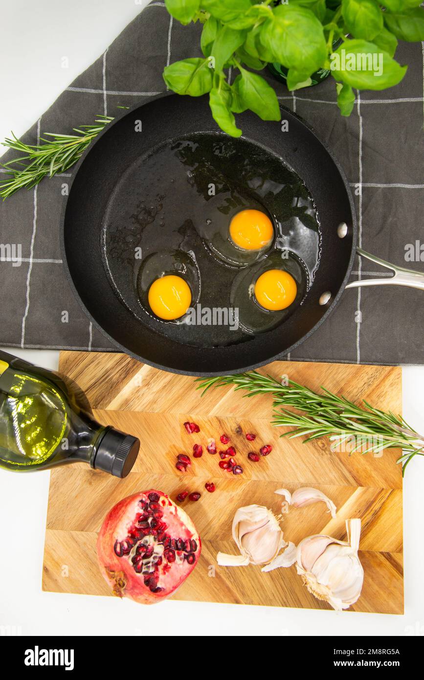 padella nera con uova crude su un tovagliolo scuro a scacchi accanto ad una tavola da cucina di legno con melograno rosso e aglio su di essa. concetto di cibo. vista dal a. Foto Stock