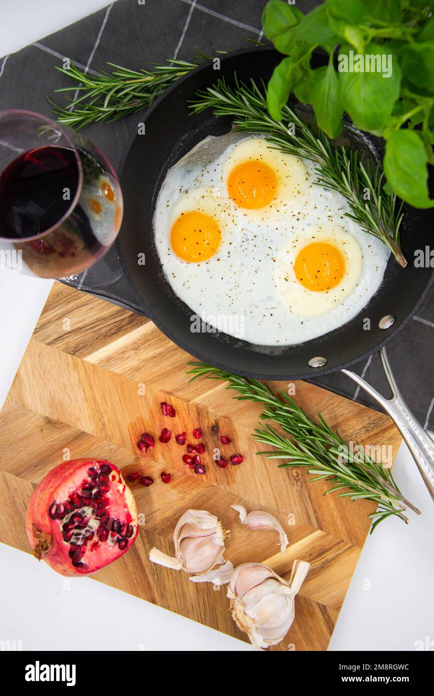 prima colazione o brunch uova fritte in una padella nera e vino rosso in un bicchiere di cristallo e melograno rosso con aglio e basilico. vista dall'alto. Foto Stock