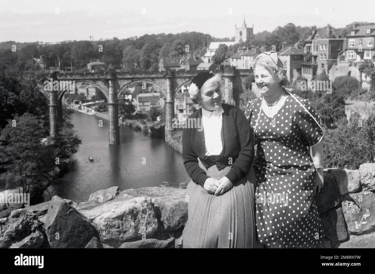 1950s, storica, due donne mature ben vestite che siedono l'una accanto all'altra sulla vecchia parete del castello a Knaresborough, North Yorkshire, Inghilterra, Regno Unito. Le rovine del castello reale si affacciano sul fiume Nidd. Un ponte sul fiume e la città di Knaresborough può essere visto sullo sfondo. Foto Stock