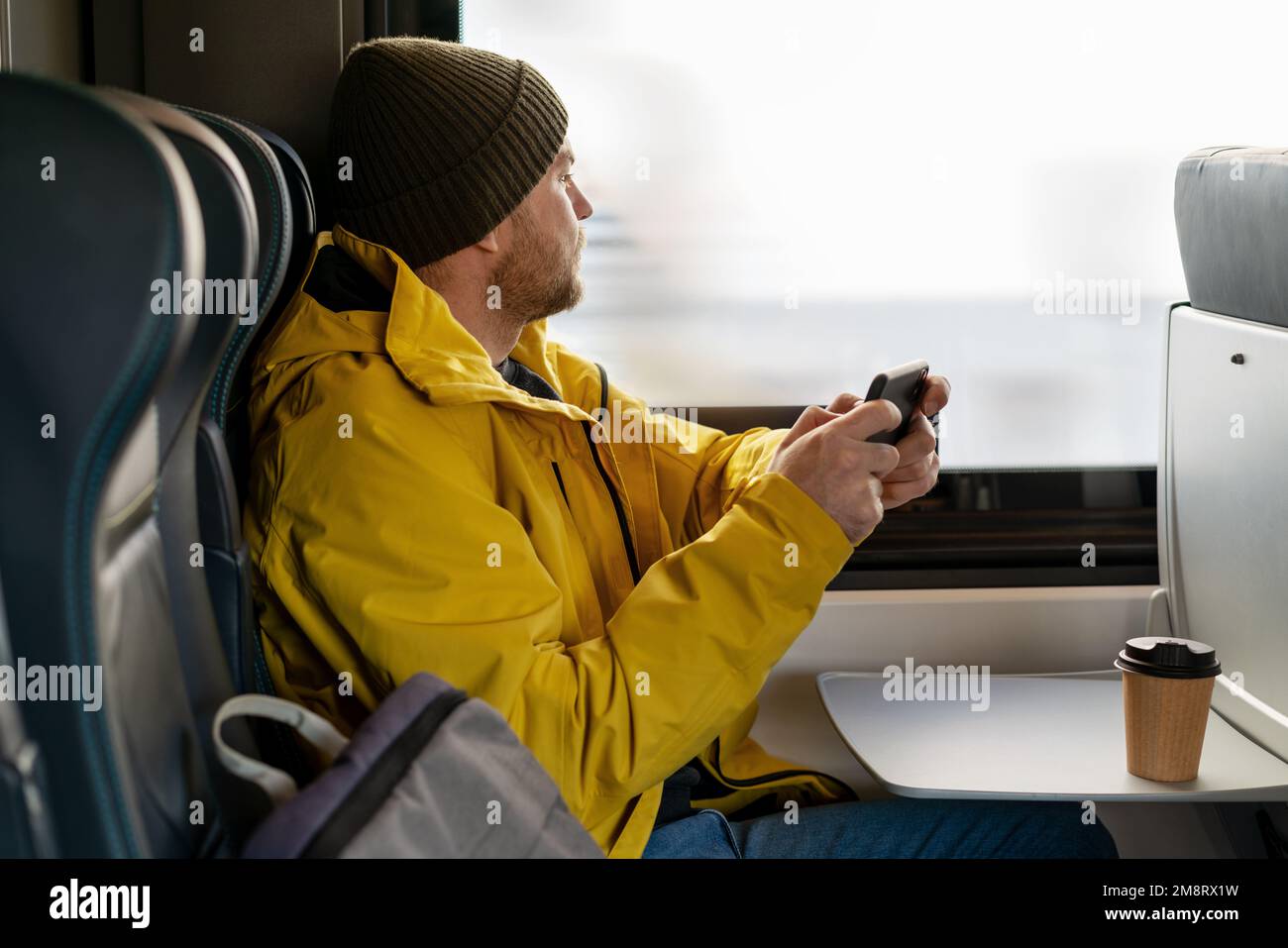 Uomo casual barbuto in giacca gialla e cappello con smartphone durante il viaggio in treno seduto all'interno del treno e guardando fuori dalla finestra. Foto Stock