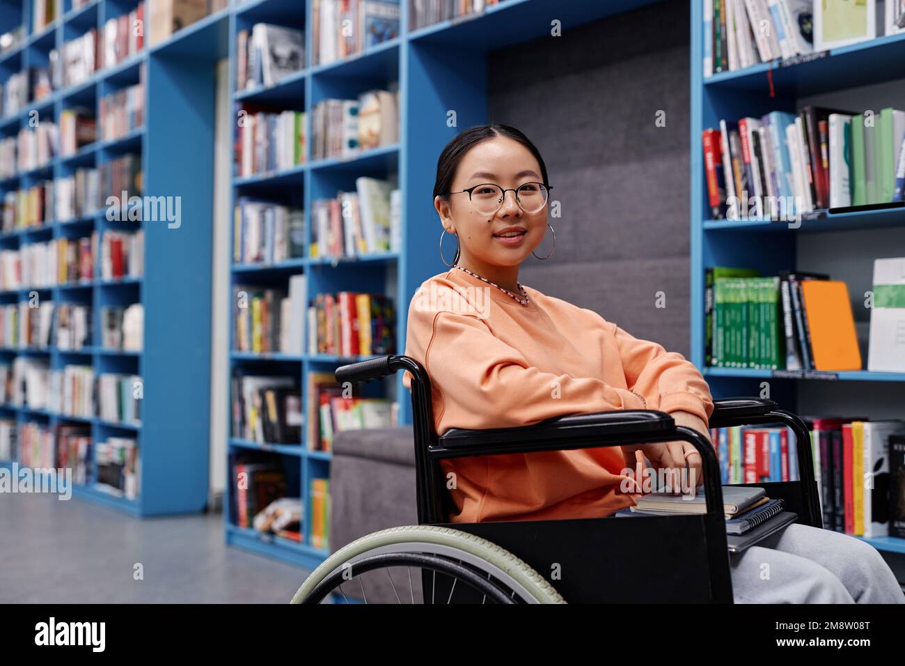 Ritratto di giovane donna asiatica con disabilità in biblioteca al college moderno sorridendo allegro, copia spazio Foto Stock