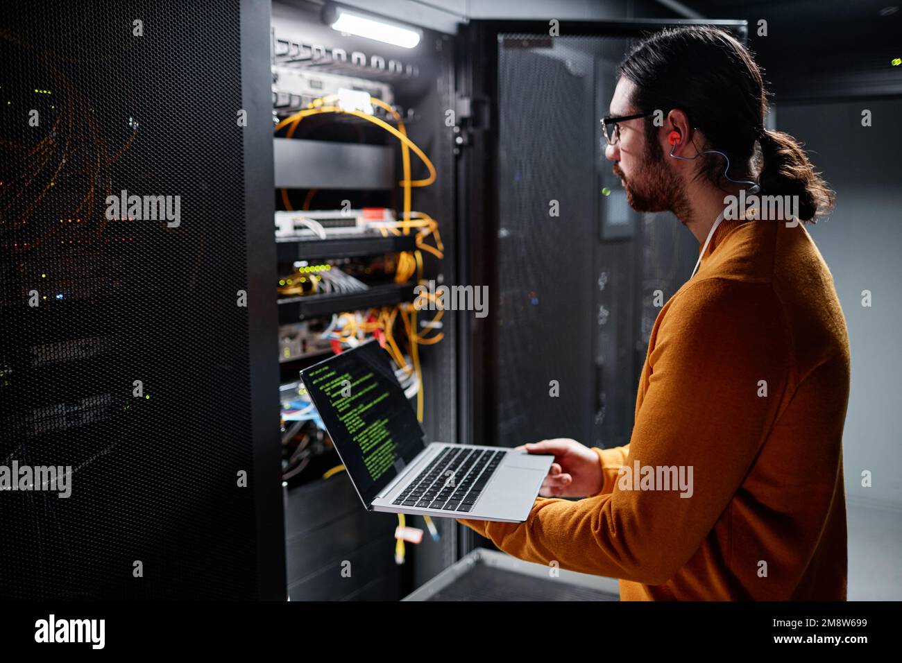Immagine laterale di un tecnico IT barbato che utilizza un notebook durante la configurazione della rete nella sala server Foto Stock