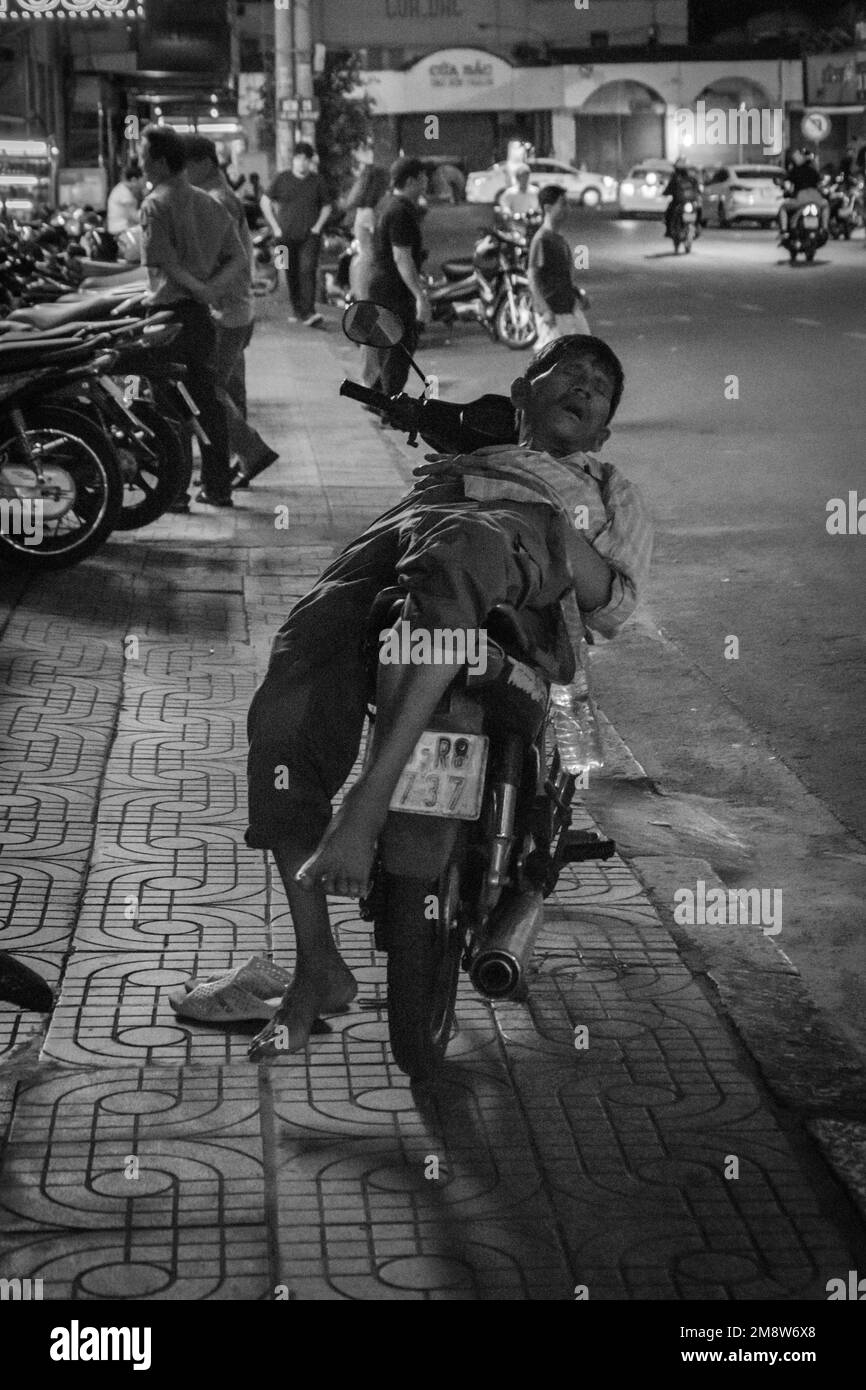 Uomo bianco e nero che dorme sulla sua moto ciclomotore per strada di notte in ho Chi Minh Vietnam Foto Stock