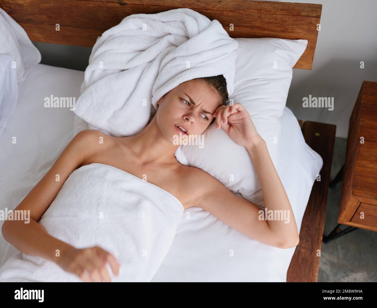 Una donna si trova su un letto al mattino su un cuscino sotto una coperta  in camera da letto dopo una doccia in un asciugamano, sentendosi malfatto  freddo e mal di testa
