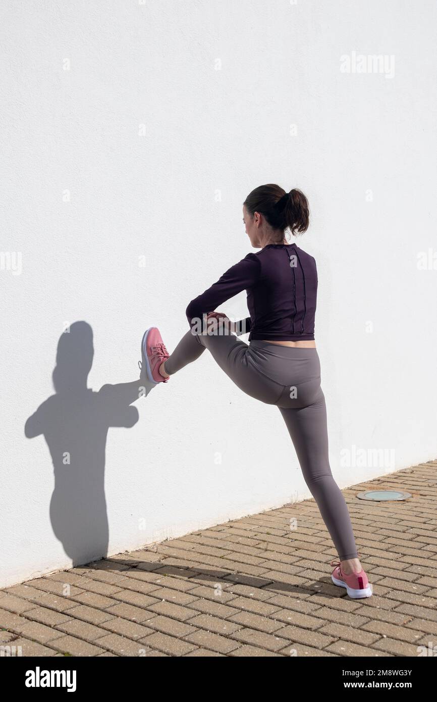 donna sportiva che fa un tratto di gamba contro una parete bianca all'esterno. Foto Stock