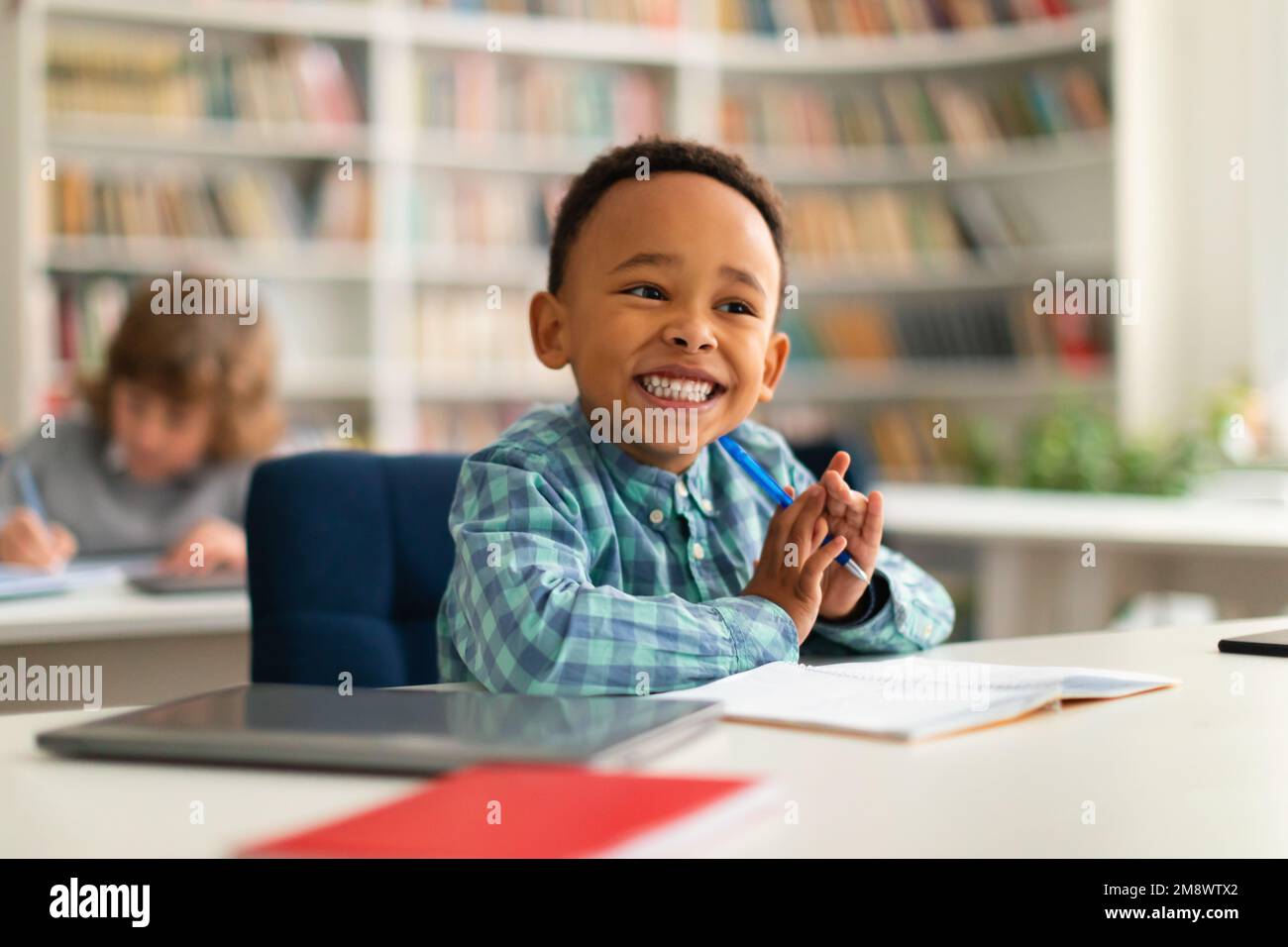 Felice ragazzo della scuola primaria afro-americana seduto alla scrivania in classe, scrivendo in notebook e sorridendo, spazio libero Foto Stock