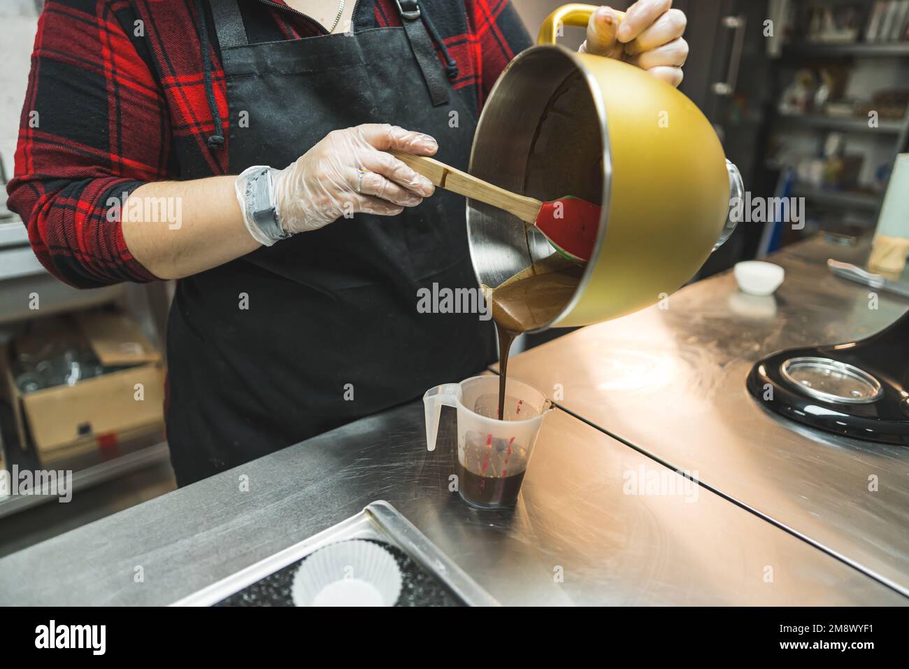 Panettiere femminile irriconoscibile con grembiule nero che raccoglie la pastella di cupcake al cioccolato nel contenitore per versarla nel vassoio di cupcake Processo di cottura professionale. Scatto orizzontale in interni. Foto di alta qualità Foto Stock