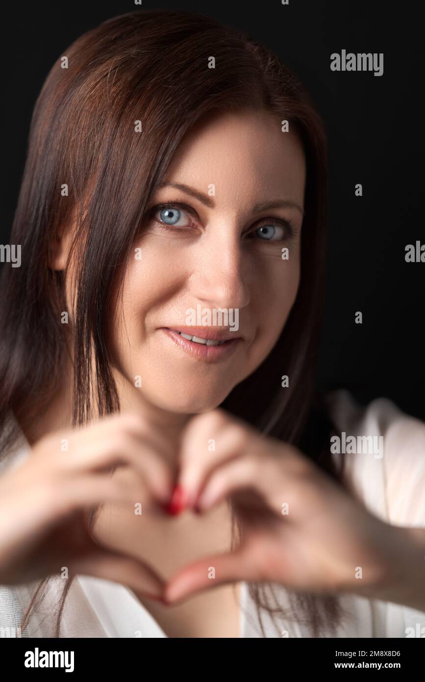 Brunette etnia caucasica donna confessa nell'amore, fa gesto cardiaco con entrambe le mani, guardando la macchina fotografica, mostrando sentimenti veri, felice espressione Foto Stock
