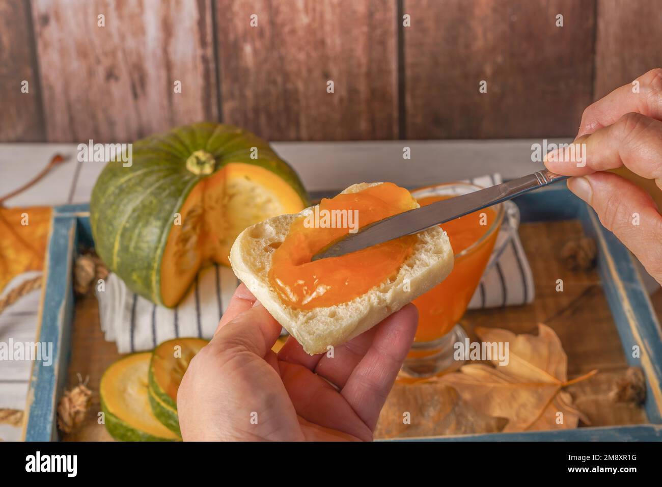 Donna che sparge marmellata di zucca fatta in casa su un pezzo di pane tostato con un coltello, alla zucca tagliata in basso e foglie secche Foto Stock