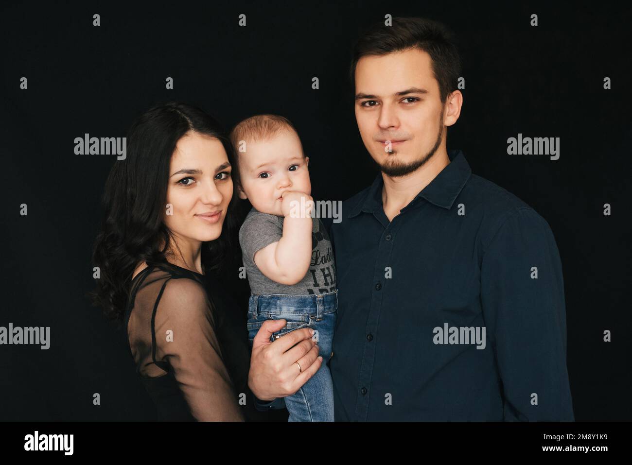 Giovane famiglia caucasica su sfondo nero nello Studio. Buon papà, mamma e figlio. Coppia amorevole che tiene un bambino. Foto Stock