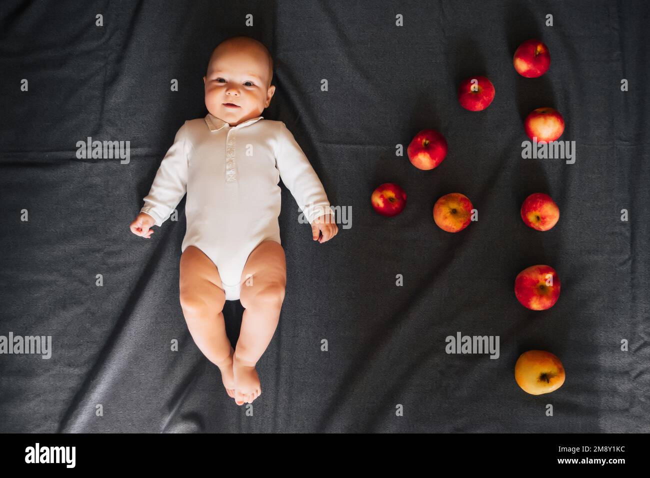 Un neonato di quattro mesi. Bambino caucasico in bodysuit bianco giacente su uno sfondo scuro con mele Foto Stock