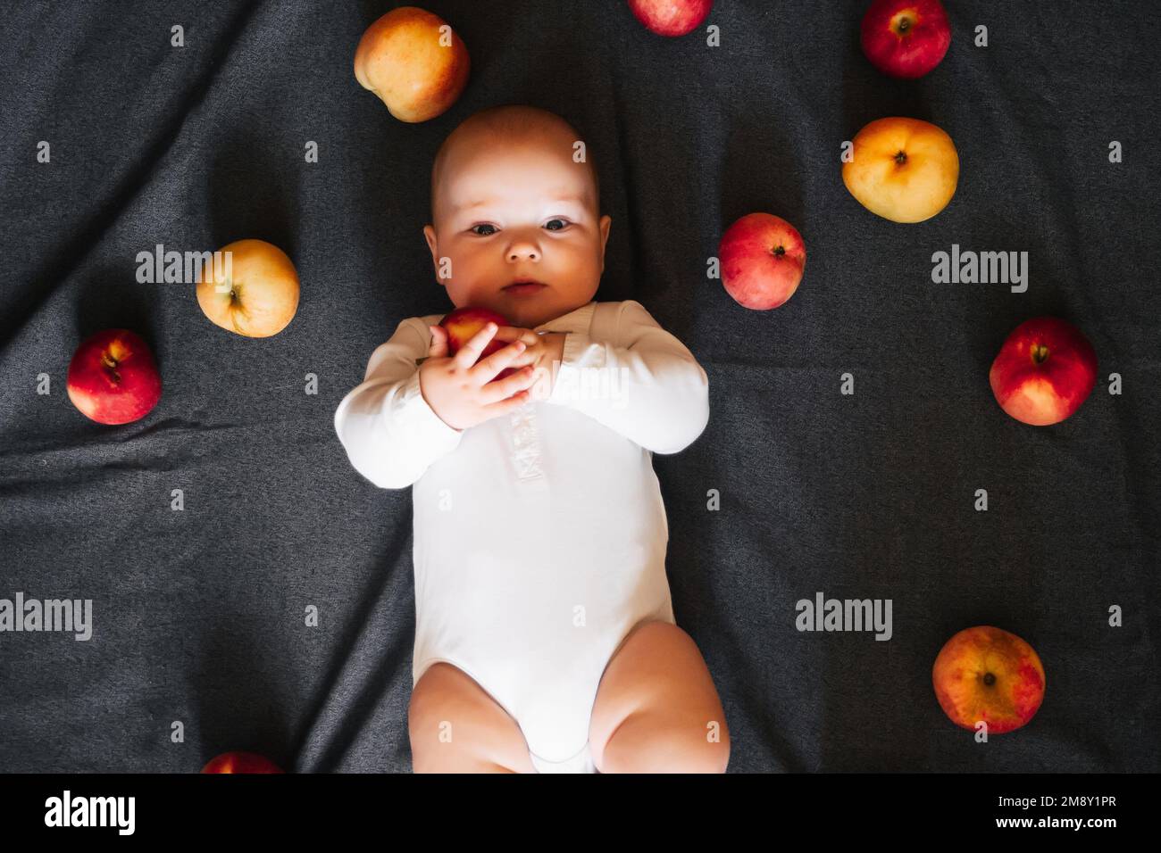 Neonato sdraiato con le mele. Un bambino piccolo in un bodysuit bianco su uno sfondo nero. Vista dall'alto, disposizione piatta Foto Stock