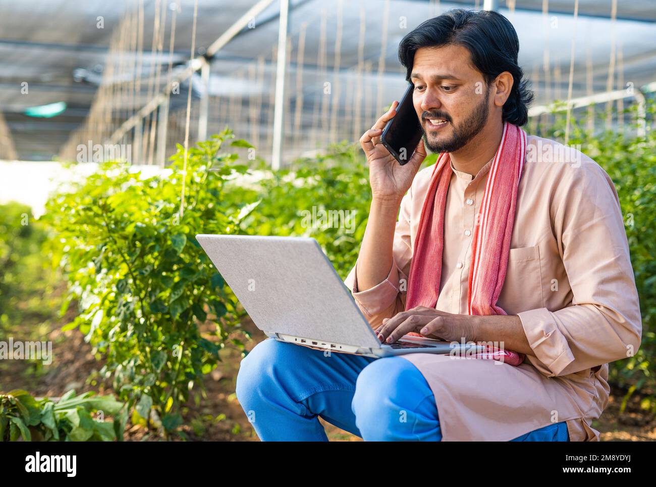 Felice agricoltore moderno a serra di lavoro o utilizzando il computer portatile parlando al telefono cellulare a serra - concetto di tecnologia, servizio clienti e. Foto Stock
