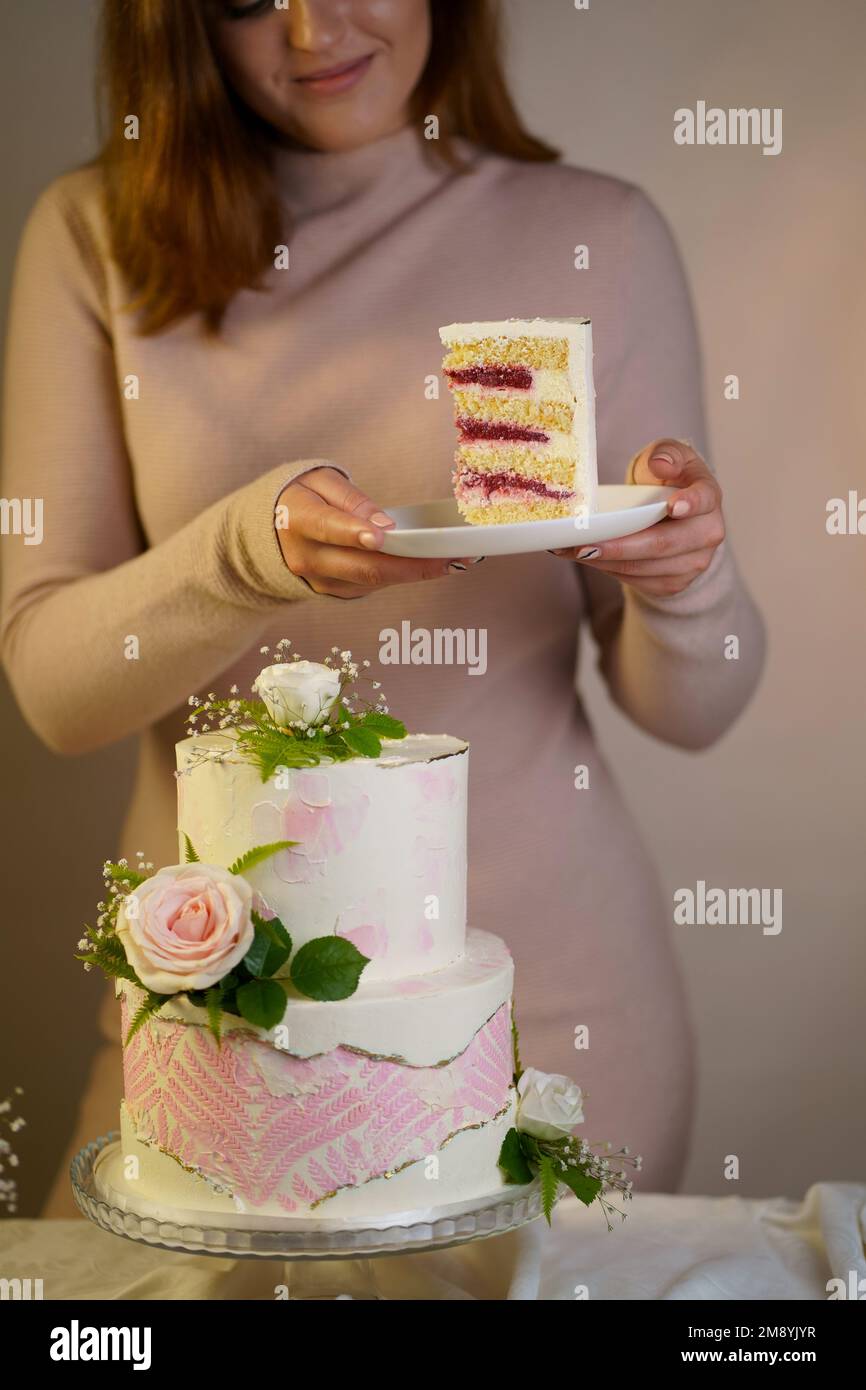 La ragazza taglia e serve un pezzo di torta. Festa di nozze torta a due livelli decorata con fiori freschi su uno sfondo grigio Foto Stock