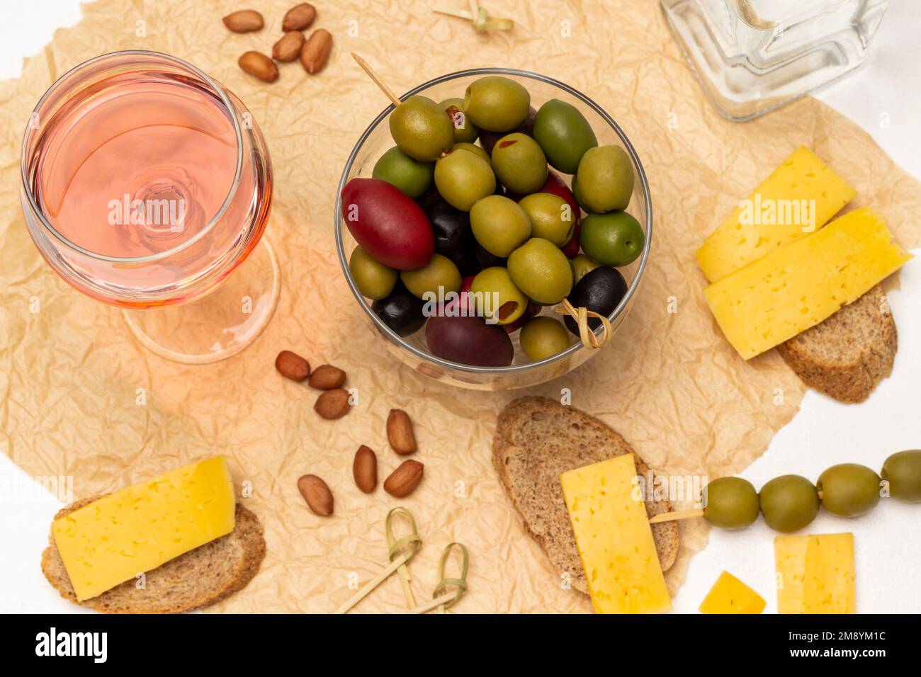Diverse varietà di olive in una ciotola di vetro. Un bicchiere di vino rosato, pane con formaggio su carta. Giacitura piatta. Sfondo bianco. Foto Stock