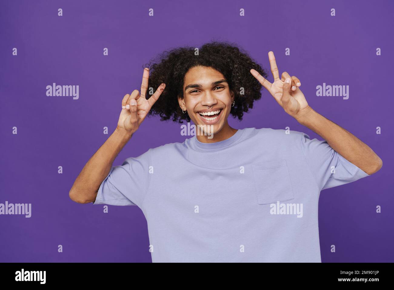 Giovane felice sorridente bell'uomo latino riccio con piercing mostrando V gesto entrambe le mani e guardando la macchina fotografica, mentre in piedi sopra isolato dorso viola Foto Stock