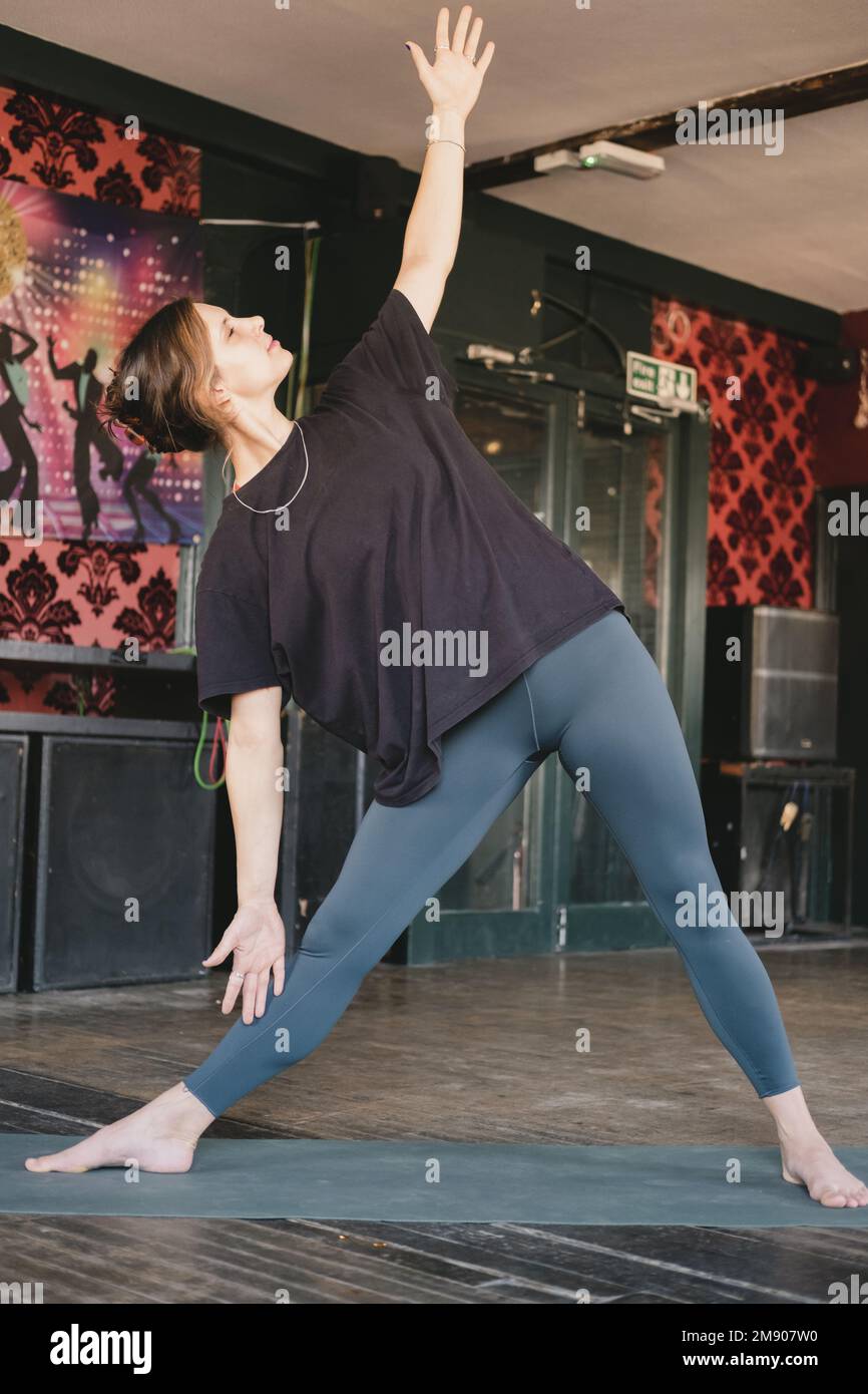 Foto verticale di giovane adulta donna yoga nuovo insegnante che fa una posa triangolare (Trikonasana) stretch, allenamento yoga, con tappetino verde e leggings Foto Stock