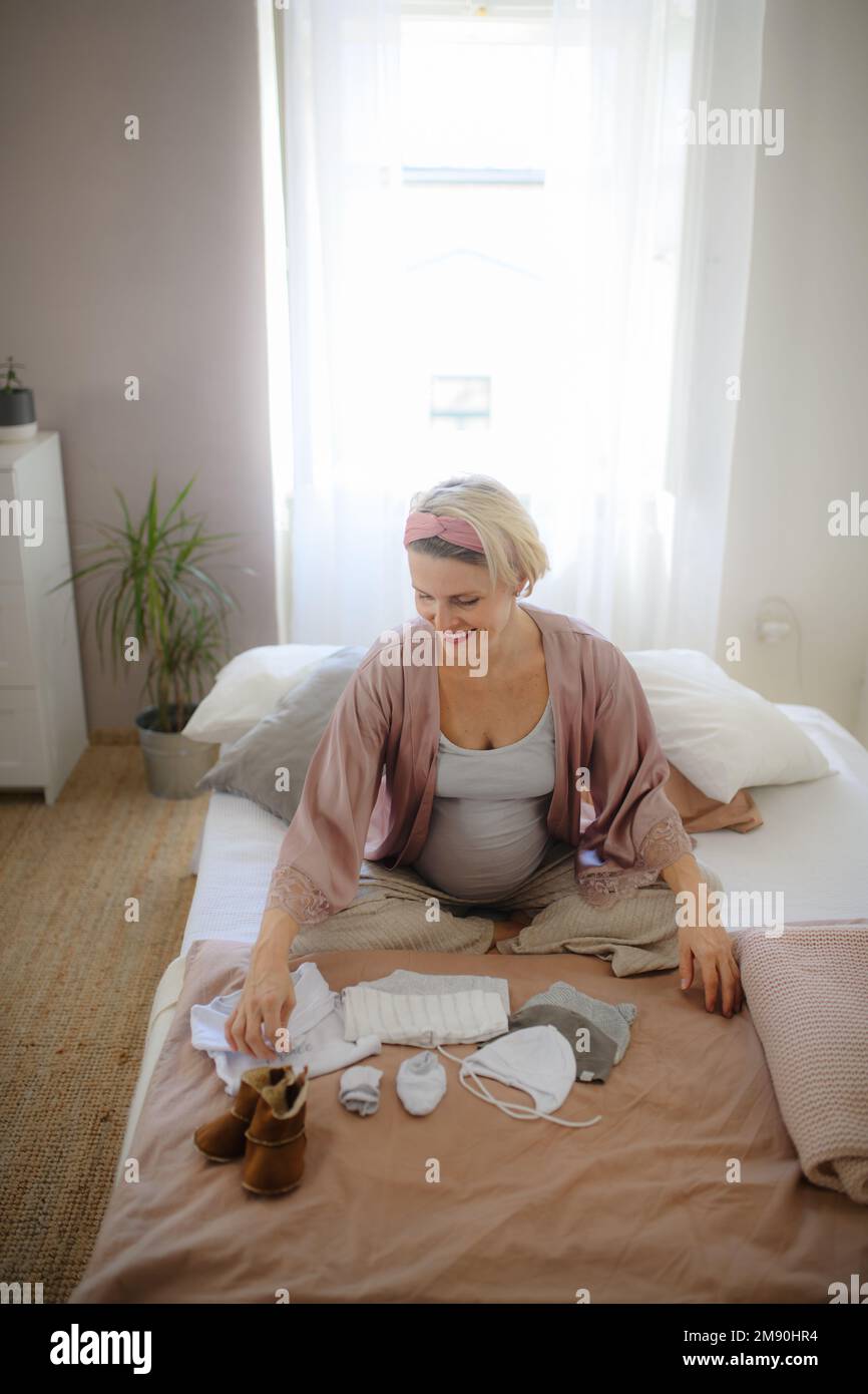 Vista ad angolo alto della donna incinta che guarda i vestiti piccoli per il suo bambino, imballandoli al reparto di maternità. Foto Stock