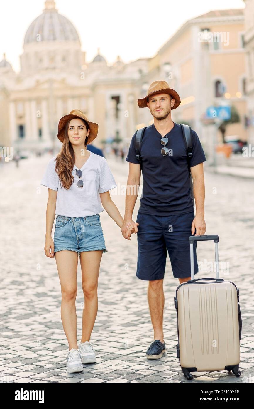 Felice giovane coppia di viaggiatori con cappelli e una valigia che abbraccia la città e godendo romanticismo. Gli amanti sorridono. Una coppia innamorata sorride e sogna Foto Stock