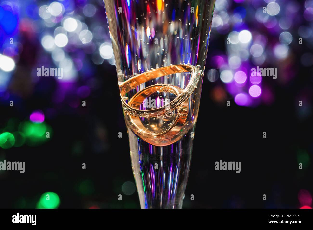 anelli di nozze d'oro in vetro di champagne con bolle d'aria su sfondo colorato sfocato primo piano Foto Stock