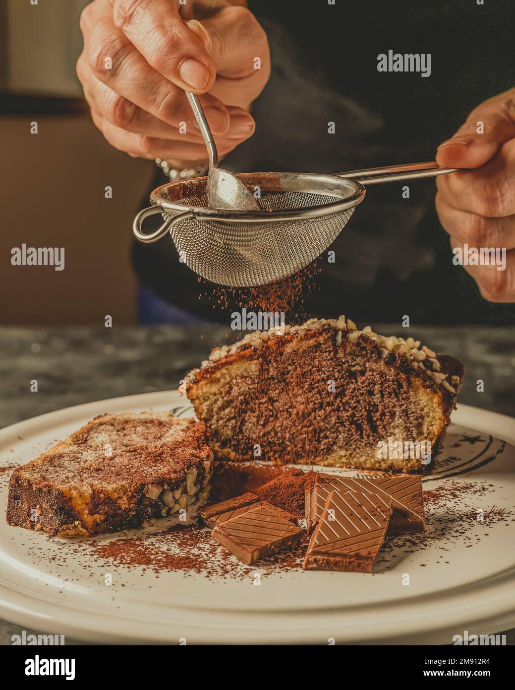 Le mani femminili tengono un setaccio dal quale la polvere di cacao cade su quadrati di cioccolato e un dessert al cioccolato e alle arachidi Foto Stock