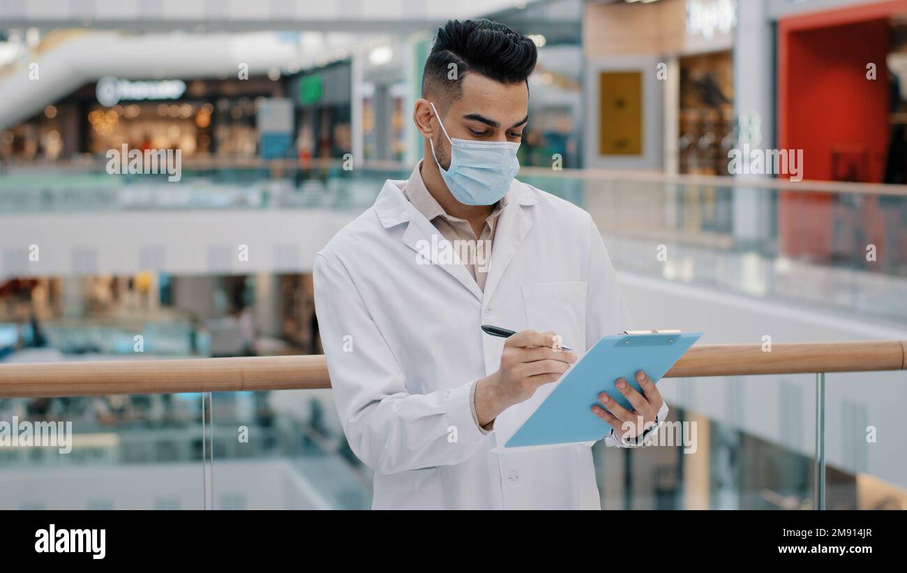 Ispettore medico indiano etnico medico scienziato medico uomo lavoratore maschio scrittura in documenti di documenti di competenza controllare la salute norme di sicurezza supervisore con Foto Stock