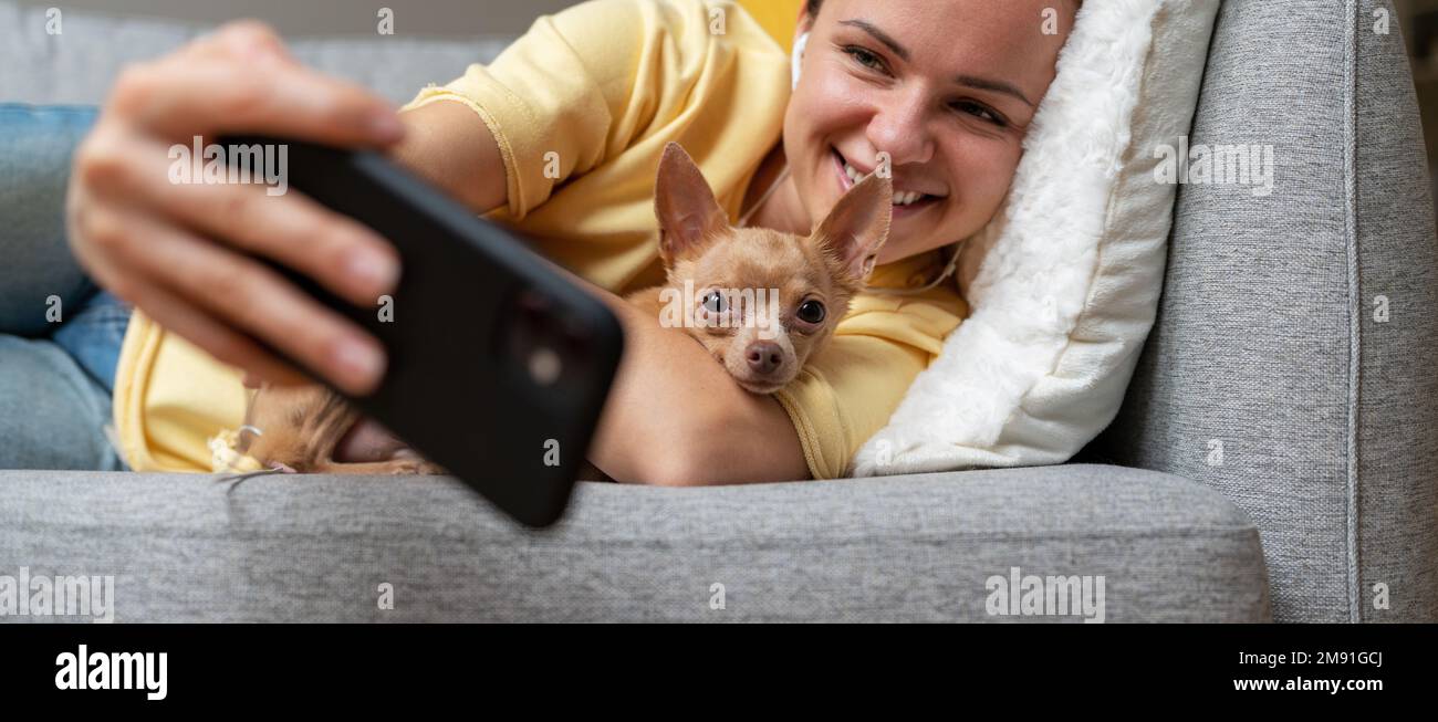 Giovane bruna donna che prende selfie utilizzando il suo smartphone mentre sdraiato sul divano con il cane piccolo. Foto Stock