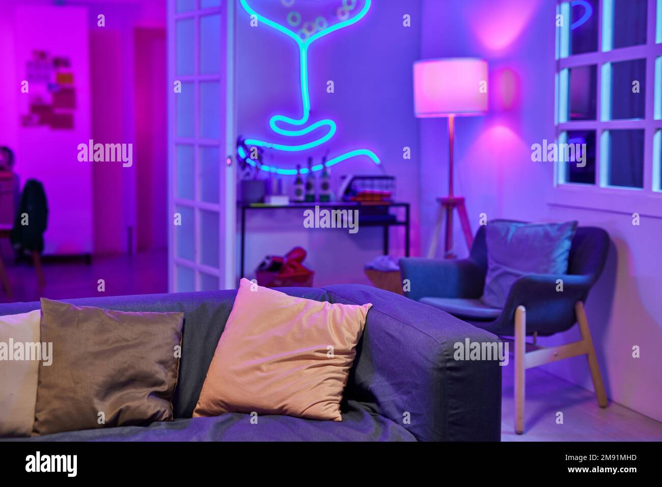 Ampio soggiorno illuminato da luce al neon con comodo divano al centro circondato da poltrona e tavolino con bevande alcoliche Foto Stock