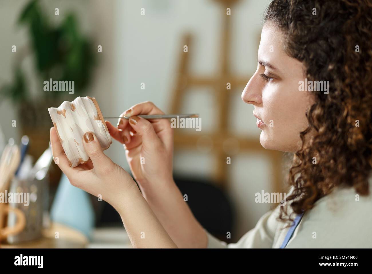 Giovane donna che fa ceramica al chiuso in studio creativo artigianale concetto seduta a tavola utilizzando strumento di modellazione creazione di dettagli sulla tazza. Foto Stock