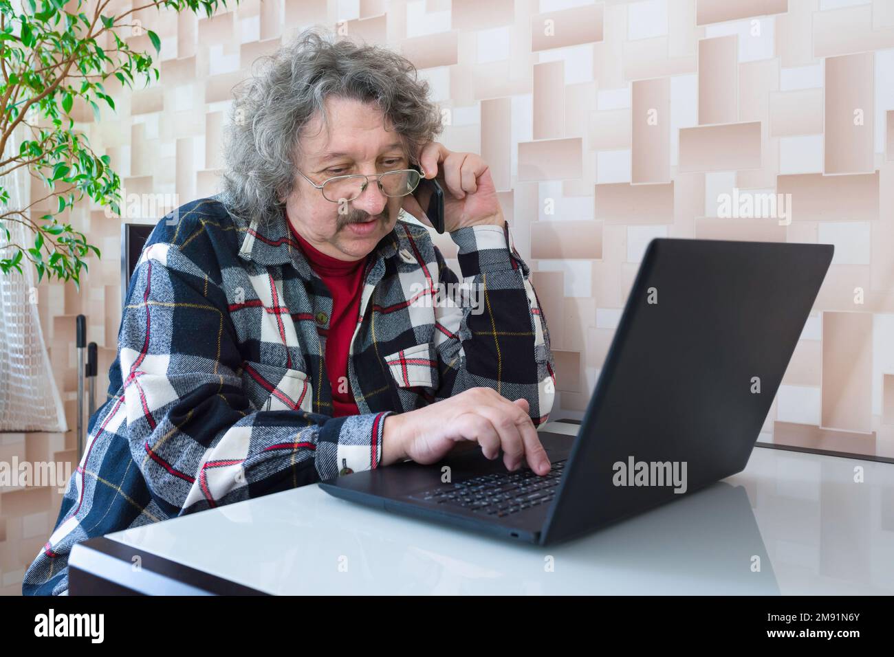 Un ritratto di un uomo anziano che lavora al computer portatile e al telefono a casa. Tecnologie, invecchiamento Foto Stock