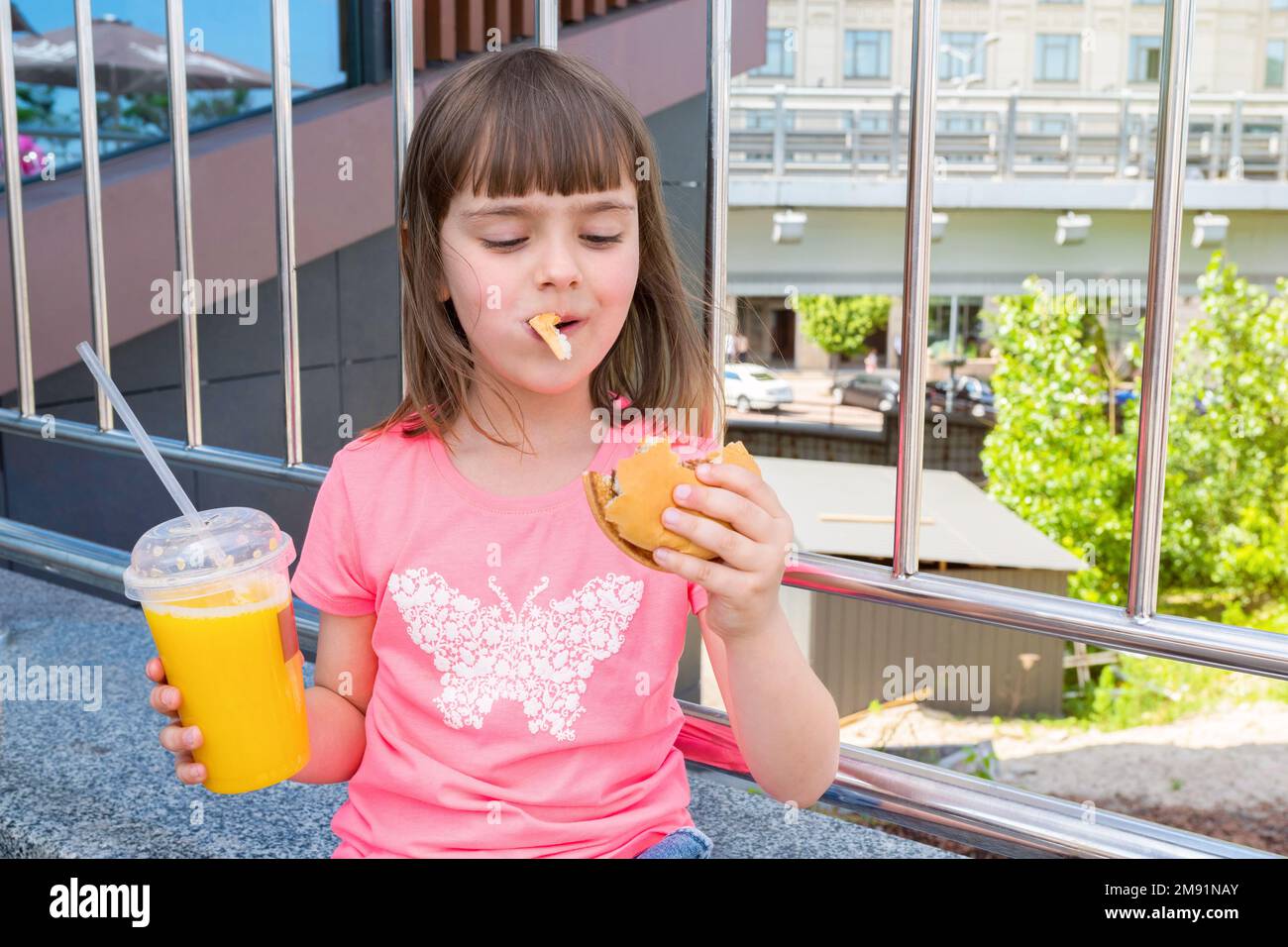 I momenti felici affamati della bella ragazza con un hamburger in bocca e con succo d'arancia. Cibo malsano, fast food grasso Foto Stock