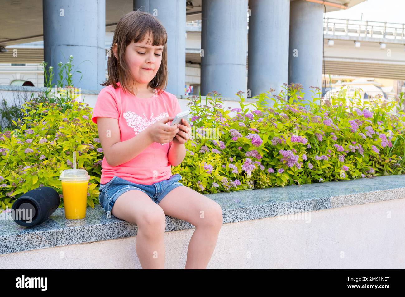 Simpatica bambina prescolare, con smartphone, seduta su un parapetto in città. Un bambino che tiene in mano uno smartphone che gioca a un gioco mobile Foto Stock