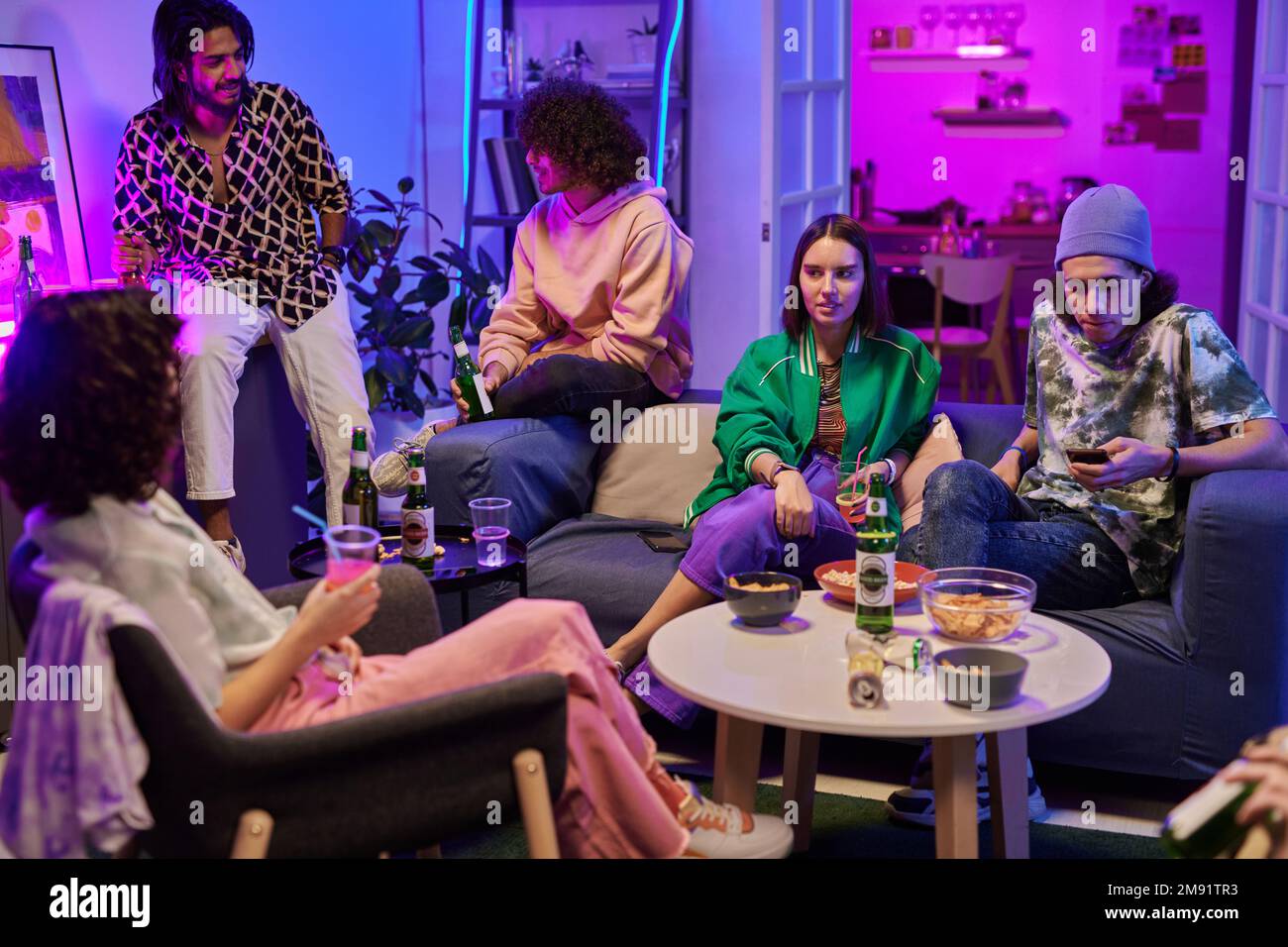 Gruppo di giovani amici in casualwear bere mentre rilassarsi nel soggiorno illuminato da luce al neon e godersi la festa a casa Foto Stock