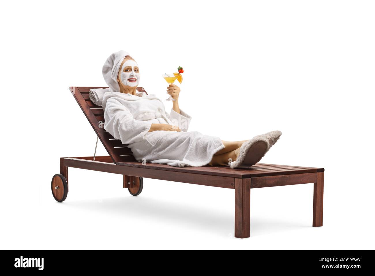 Donna in accappatoio che indossa una maschera anti-invecchiamento e che tiene un bicchiere di cocktail isolato su sfondo bianco Foto Stock