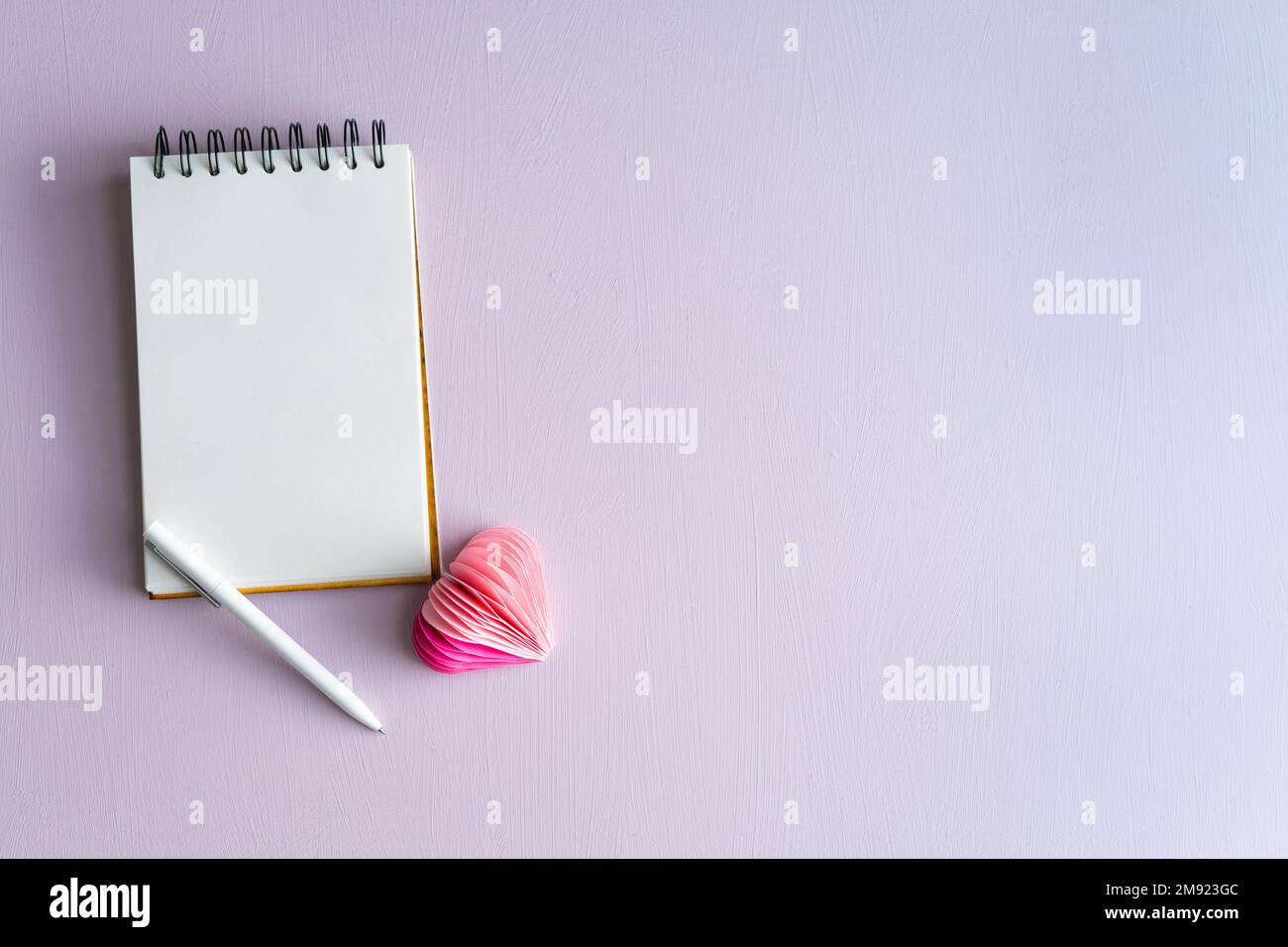 Notebook vuoto con penna e cuore di carta su sfondo lilla. San Valentino e Festa della mamma. Spazio di copia. Foto Stock