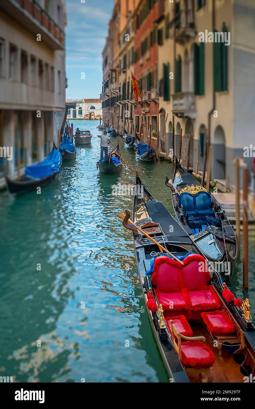 Gondoliere veneziane che naviga attraverso un canale laterale più piccolo verso il Canal Grande, passando per diverse bellissime gondole Foto Stock