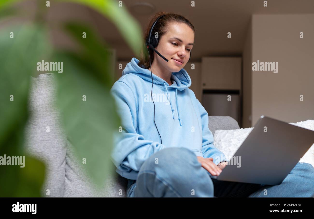 Donna bruna in cuffia seduta sul divano a casa e che utilizza un computer portatile che lavora a casa e-learning a distanza. Foto Stock