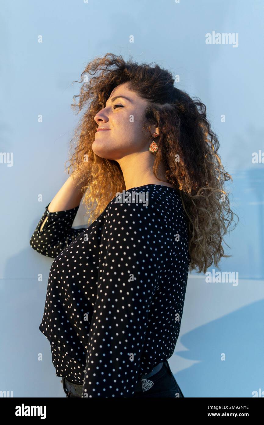 Vista laterale angolo basso di giovane donna con capelli ricci in blusa pois polka in piedi contro la parete blu nei giorni di sole che toccano i capelli Foto Stock