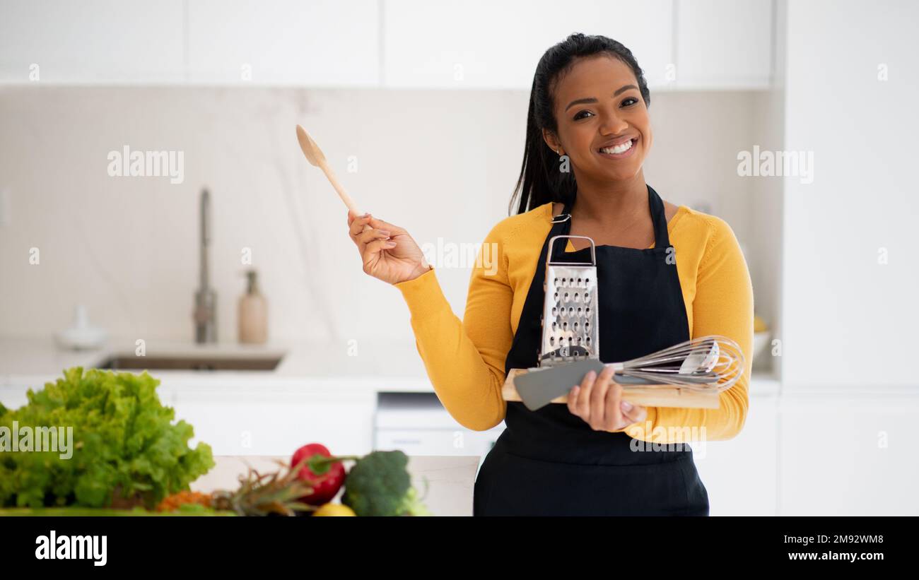Giovane ragazza afro-americana sorridente in grembiule con cucchiaio di legno, grattugia e frusta Foto Stock