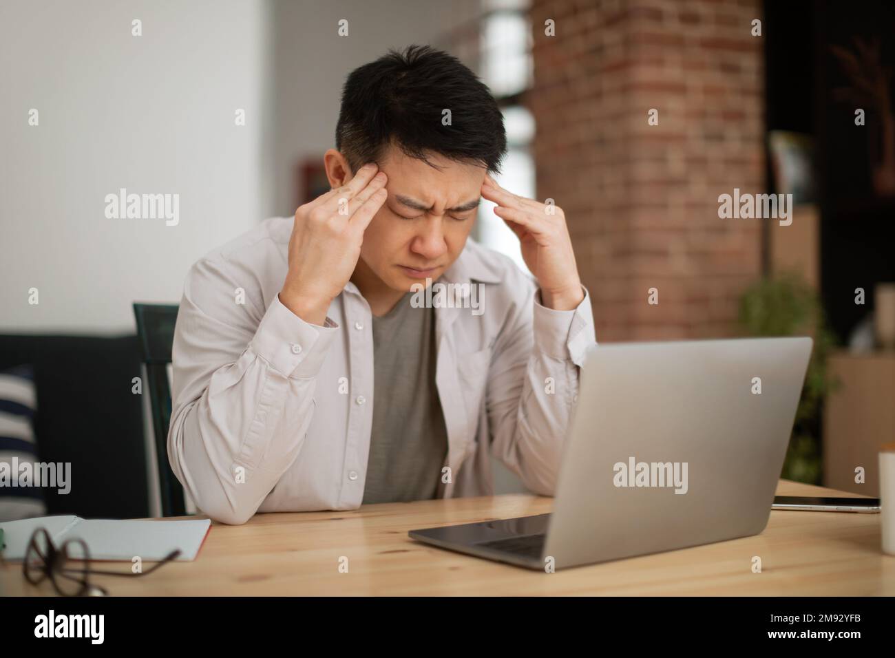 Triste maturo libero professionista asiatico che lavora su un computer portatile e soffre di mal di testa, seduto al tavolo in ufficio a casa Foto Stock