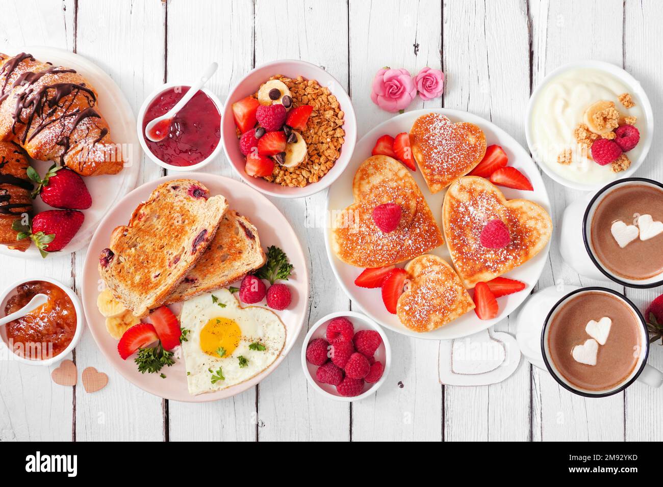 Tavolo per brunch di San Valentino o Mothers Day. Vista dall'alto su uno sfondo di legno bianco. Frittelle a forma di cuore, uova e un assortimento di piatti a tema d'amore. Foto Stock