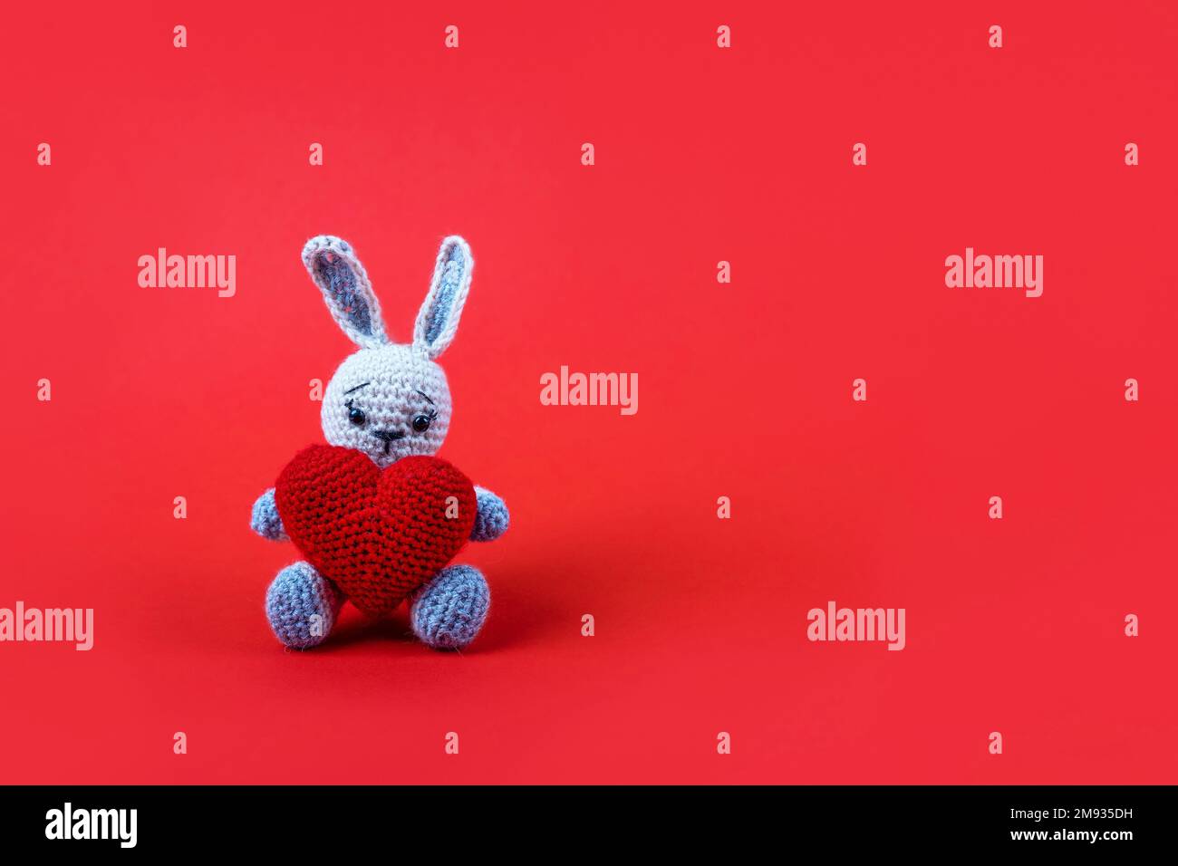 Coniglietto grigio lavorato a maglia con cuore rosso uncinetto su sfondo rosso. Buon San Valentino, festa della mamma e auguri di compleanno. Foto Stock