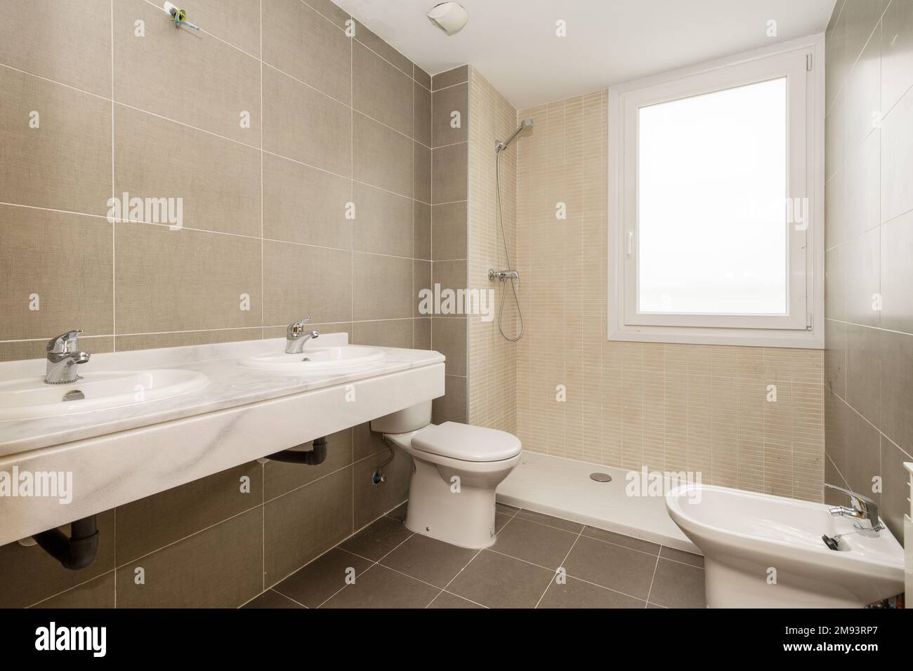 Nuovo bagno senza specchio e divisorio dotato di WC in porcellana bianca e piastrelle grigie e crema, finestra in doccia e doppio lavabo su marmo c Foto Stock