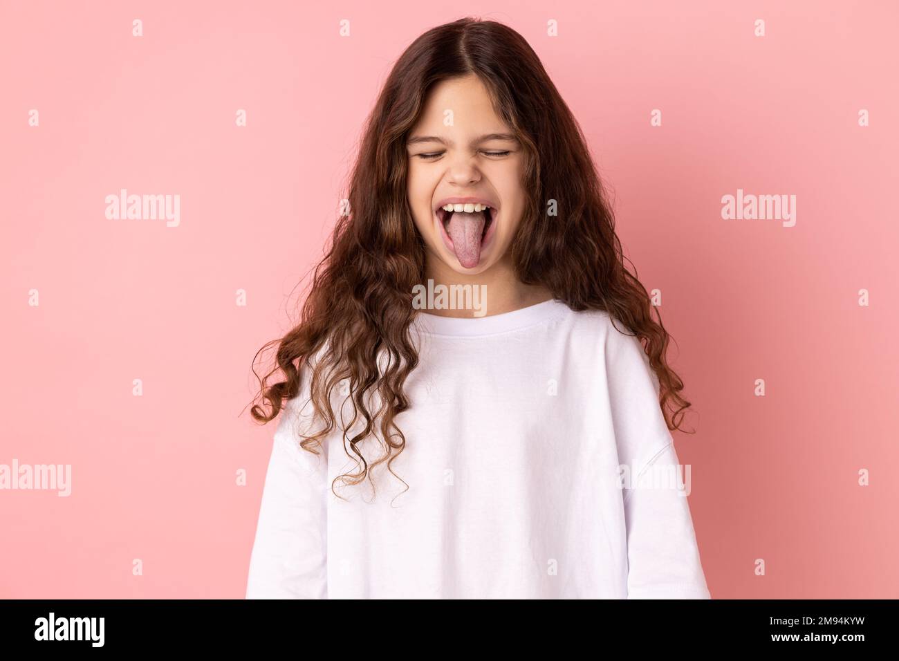 Ritratto di bambina spensierata e infantile che indossa una T-shirt bianca che mostra la lingua e chiude gli occhi con una grimace disubbidiente, facendo il viso. Studio in interni isolato su sfondo rosa. Foto Stock