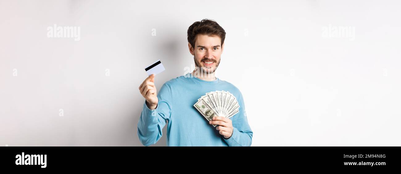 Bel giovane uomo sorridente e che offre il pagamento in contanti e senza contatto, mostrando denaro con carta di credito in plastica, in piedi su sfondo bianco Foto Stock