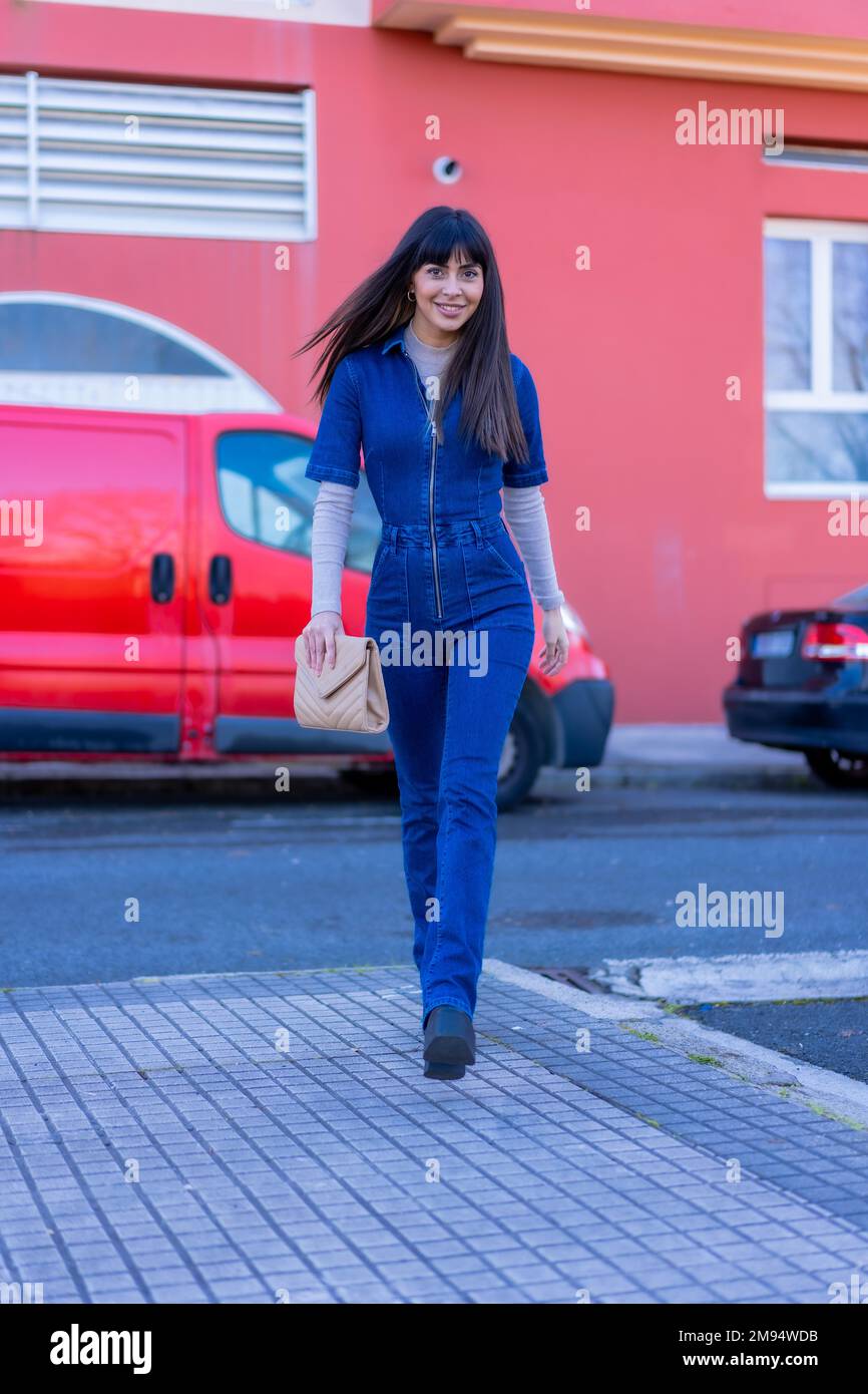 Posa di un modello di brunetta che cammina per la strada con un abito in denim. Caucasico piuttosto uno sfondo di case rosse Foto Stock