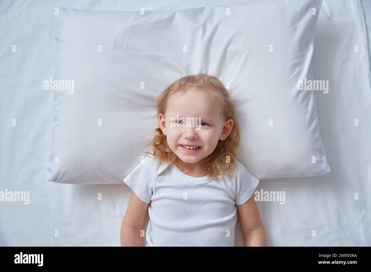 Vista dall'alto testa bambina di bambina dai capelli ricci sdraiata letto lenzuolo bianco, cuscino. Beffa per l'immagine di corna di cervo. Il concetto di buon umore e Capodanno Foto Stock