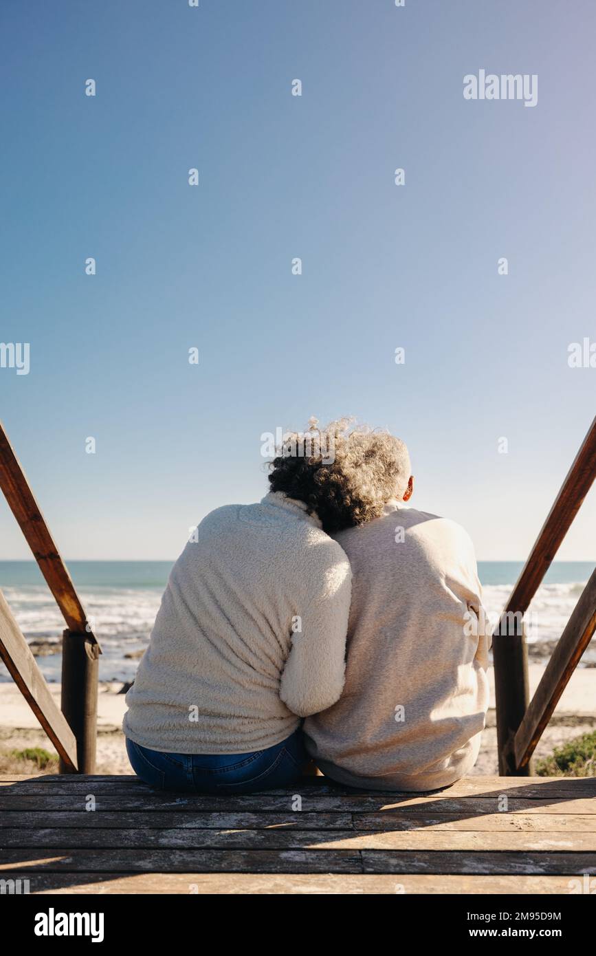 Vista posteriore di una coppia anziana che ha una rinfrescante vista dell'oceano mentre si siede su un ponte pedonale di legno. Coppia anziana in pensione che spende qualche quali Foto Stock