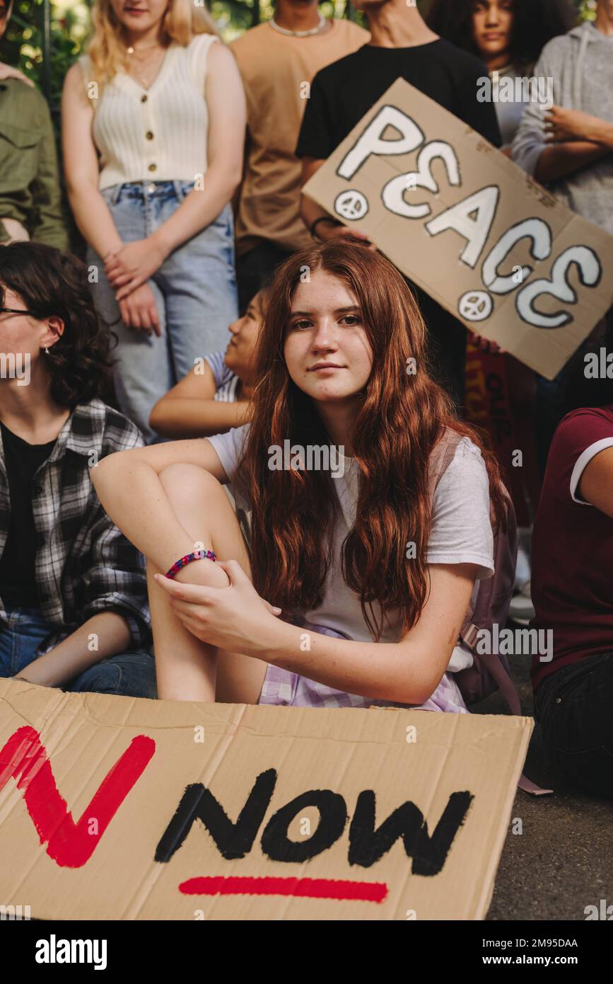 Giovane ragazza che guarda la telecamera mentre siede con un gruppo di attivisti della pace giovanile. Giovani multietnici che espongono poster e banner mentre pr Foto Stock