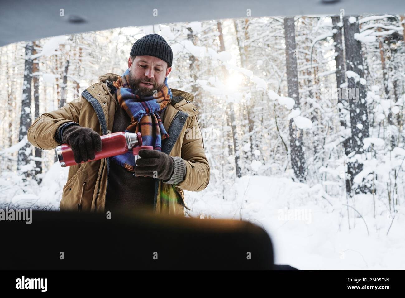 Giovane uomo bearded in abiti caldi bere tè caldo durante il pic-nic nella foresta d'inverno Foto Stock
